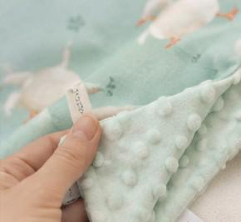 May include: A light mint green baby blanket featuring a repeating pattern of white geese. The fabric has a textured dimpled surface on one side and a smooth side with the printed geese. A small white fabric tag with black text is attached to the edge.