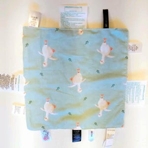 May include: A square light blue fabric square featuring a repeating pattern of white geese and small green leaves. The fabric is surrounded by multiple white and black fabric care and content labels attached to all four edges of the square.
