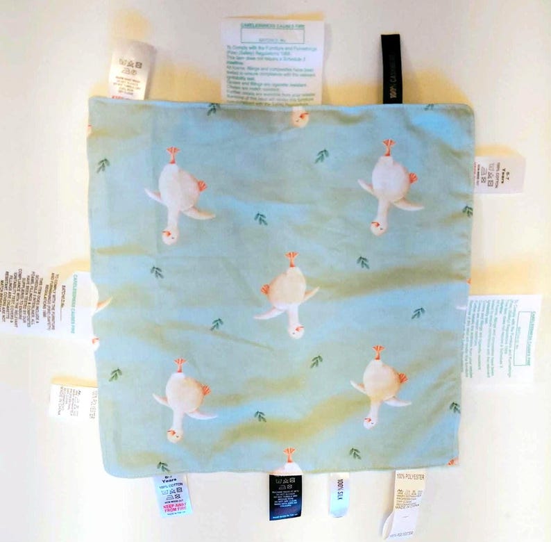 May include: A square light blue fabric square featuring a repeating pattern of white geese and small green leaves. The fabric is surrounded by multiple white and black fabric care and content labels attached to all four edges of the square.