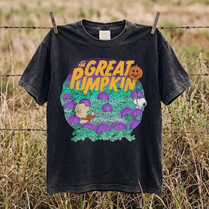 May include: A black short-sleeve t-shirt featuring a graphic print of Charlie Brown and Snoopy in a pumpkin patch. The text reads IT'S THE GREAT PUMPKIN in yellow letters. The design includes purple pumpkins and green foliage on a dark background.