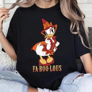 May include: A black t-shirt featuring a graphic of a white duck character wearing an orange and red witch hat and cape. The character has yellow feet and is posed with one hand near its chin. Below the graphic, the text FA-BOO-LOUS is printed.