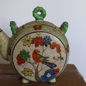 May include: A green and white ceramic teapot with a floral design and a bird. The teapot has a lid with a green knob and a spout. The teapot is decorated with red, yellow, blue, and green flowers and a bird.