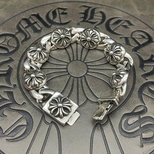 May include: A silver bracelet with a floral design. The bracelet features a series of flower-shaped links connected by a chain. The clasp is also adorned with a flower design. The bracelet is displayed on a gray surface.
