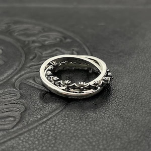 May include: A silver ring with a three-band design. The outer bands are smooth, while the inner band features a repeating floral pattern. The ring is displayed on a dark, textured surface.