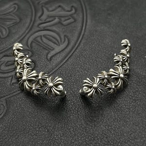 May include: Two silver ear cuffs, each featuring a series of flower-shaped designs. The cuffs curve to fit the ear, offering a distinctive look. The jewellery is presented on a dark, textured surface, highlighting the intricate details of the design.