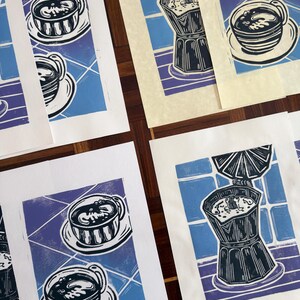 May include: Multiple rectangular art prints featuring linocut-style illustrations of moka pots and coffee cups on a blue tiled background. The prints use a color palette of dark blue, light blue, and white, showcasing coffee brewing themes.