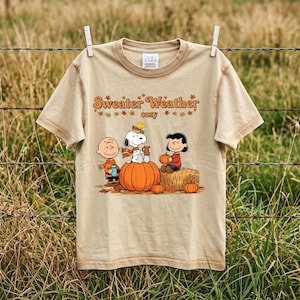 May include: A beige short-sleeve t-shirt featuring a graphic print of Charlie Brown, Snoopy, Woodstock, and Lucy sitting on pumpkins. The text "Sweater Weather" and "cozy" is printed above the characters, surrounded by small orange autumn leaves.
