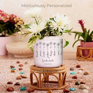 May include: A white personalized flower pot with the text "Grandma Linda's Garden Of Love" and names. The pot is filled with white flowers and sits on a small, round, woven stand. The words "A Gift to Grow On" are also visible.