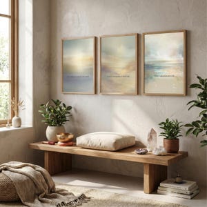 May include: Three framed rectangular wall art prints featuring abstract, soft-toned landscapes in muted shades of blue, beige, and pink. Each print contains small, centred text. The artwork is displayed in a row on a textured, light-coloured wall.