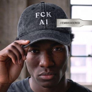 May include: A charcoal grey acid-wash baseball cap featuring white embroidered text on the front that reads FCK AI. The cap has a curved brim and a structured crown with a visible //EMBROIDERED tag attached to the side of the fabric.