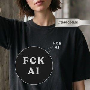 May include: A black short-sleeved t-shirt featuring white embroidered text on the upper left chest that reads FCK AI. The embroidery has a textured appearance, and a circular inset highlights the detail of the stitched lettering on the fabric.