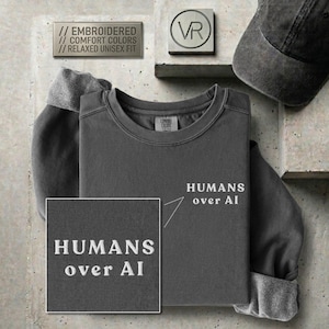 Puede incluir: Una sudadera de cuello redondo en color gris carbón con el texto blanco bordado "HUMANS over AI" en el pecho. La prenda está confeccionada en un tejido suave y relajado. Una gorra de béisbol a juego en gris carbón se sitúa cerca.