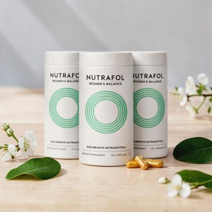Pode incluir: Três frascos cilíndricos brancos de Nutrafol Women's Balance, um suplemento para o crescimento capilar. Cada frasco tem um design de círculos concêntricos verdes e o texto "Hair Growth Nutraceutical" e "120 Capsules". Várias cápsulas douradas estão em frente aos frascos.