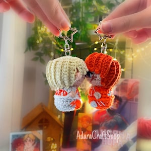 May include: Two handmade crochet keychains, one with blonde hair and the other with red hair, each with a star-shaped clasp. The keychains are in the shape of small figures, with details in orange, white, and gray.