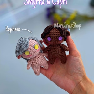 May include: Two handmade crochet doll keychains, one pink and gray, the other brown. The pink doll has yellow eyes and gray hair, while the brown doll has purple eyes and brown hair. The text "Smyrna & Capri" and "Keychains" are visible.