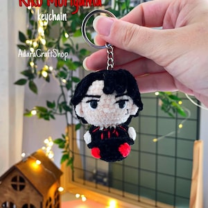 May include: A crochet keychain featuring a character with black hair, a pale face, and black clothing with red accents. The figure has embroidered eyes and a small mouth. The text Riko Moriyama keychain and AdaraCraftShop is visible in the upper left corner.
