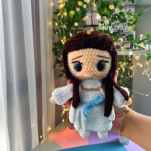 Puede incluir: Una muñeca de ganchillo hecha a mano con pelo de lana marrón oscuro, ojos negros grandes y cara de color melocotón claro. La muñeca viste una túnica blanca texturizada con un cinturón de lana azul claro y un collar de lana naranja fino.