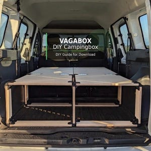 Puede incluir: Una caja de camping de bricolaje, construida con madera de color claro, está instalada en la parte trasera de una furgoneta blanca. La caja es rectangular con bisagras y soportes negros. El texto en la imagen dice "VAGABOX DIY Campingbox DIY Guide for Download."