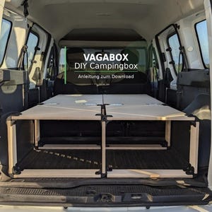 Puede incluir: Caja de camping de madera de color claro instalada en la parte trasera de una furgoneta blanca. La caja es rectangular con bisagras y soportes negros. El texto en la imagen dice "VAGABOX DIY Campingbox".