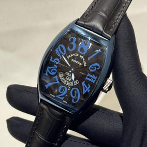 May include: A luxury wristwatch with a black face and blue numerals. The watch has a blue case and a black leather band. The watch face displays the brand name "FRANCK MULLER GENEVE" and the words "BLACK BADGE".