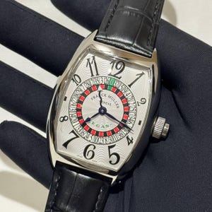 May include: A silver wristwatch with a black leather strap. The watch face has a roulette wheel design with black and red numbers. The brand name "FRANCK MULLER GENEVE" is displayed. The watch has a silver case and a textured dial.