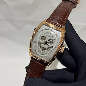 May include: A luxury wristwatch with a skull-shaped face. The watch has a gold-toned case and a brown leather strap. The face is silver with the words "FRANCK MULLER GENEVE" and a skull design.
