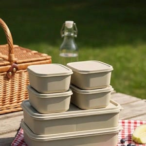Puede incluir: Un conjunto de recipientes cuadrados beige para guardar alimentos con tapas, apilados sobre una superficie de madera. En el fondo, una cesta de picnic, una botella de vidrio y un paño a cuadros rojos y blancos. Arándanos y rodajas de pepino en primer plano.