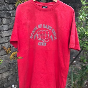 May include: A red t-shirt with the text "SCHOOL OF HARD KNOX" in gray, featuring a fist graphic with the word "FUCT" inside. The shirt has a crew neck and short sleeves. The brand name "FUCT" is printed on the collar.