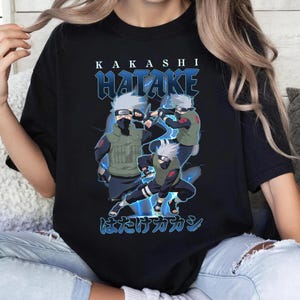 May include: A black short-sleeve t-shirt featuring a graphic print of the anime character Kakashi Hatake in three different poses. The design includes the text KAKASHI HATAKE in white and blue, with blue lightning accents and Japanese characters.