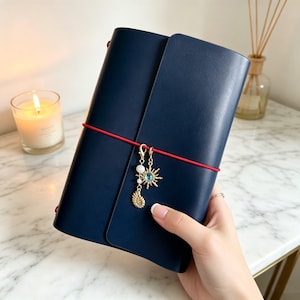 May include: A navy blue leather journal with a red elastic closure. The journal is adorned with a gold charm featuring a sun, shell, and beads. The journal is held in a hand.