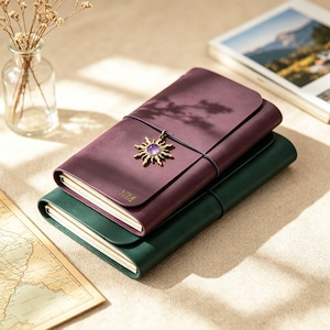 May include: Two leather journals, one deep purple and one forest green, stacked on a beige surface. The purple journal has a sun-shaped charm with a purple stone and the initials "NZ" embossed. A small glass vase with dried flowers is in the background.