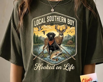 May include: An olive green t-shirt featuring a graphic print with the text "LOCAL SOUTHERN BOY" and "Hooked on Life". The design depicts a black Labrador wearing a red bandana, a deer, a jumping fish, fishing rods, and a landscape scene.