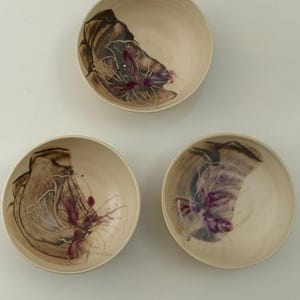 Op de afbeelding: Drie ronde keramische kommen met een crèmekleurige basis. Elke kom heeft een interieurontwerp bestaande uit een bruine, penseelstreekachtige vlek, geaccentueerd met dunne witte lijnen en een levendig magenta bloem- of abstract patroon.