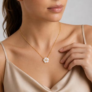 Peut inclure: Un collier à fine chaîne dorée orné d'un pendentif en forme de fleur blanche à cinq pétales arrondis et un petit centre doré. Le collier repose sur la clavicule, mettant en valeur une fine chaîne en métal poli avec des perles dorées espacées.