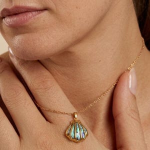Peut inclure: Un collier doré avec un pendentif en forme de coquillage. Le pendentif présente une monture en métal doré avec des stries verticales et une incrustation en nacre d'ormeau irisée aux reflets multicolores. La chaîne est en maillons dorés.
