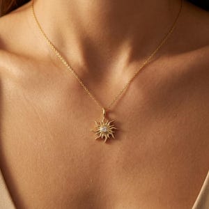 Puede incluir: Un collar de tono dorado con un colgante en forma de sol que presenta una piedra preciosa redonda y transparente en el centro. El colgante cuelga de una delicada cadena dorada que descansa sobre la clavícula, mostrando un diseño radiante.