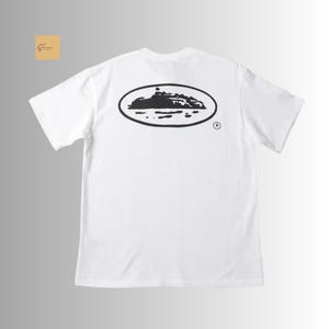 May include: A white short-sleeve t-shirt featuring a black graphic on the back. The graphic consists of an oval outline containing a silhouette of a landmass or island surrounded by water, with a small registered trademark symbol to the right.