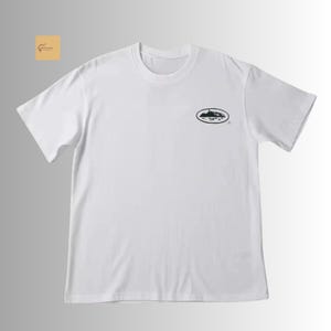 May include: A white short-sleeved t-shirt featuring a crew neckline and a small black oval graphic on the upper left chest. The graphic depicts a detailed landscape illustration inside the oval shape, positioned neatly against the plain white fabric.