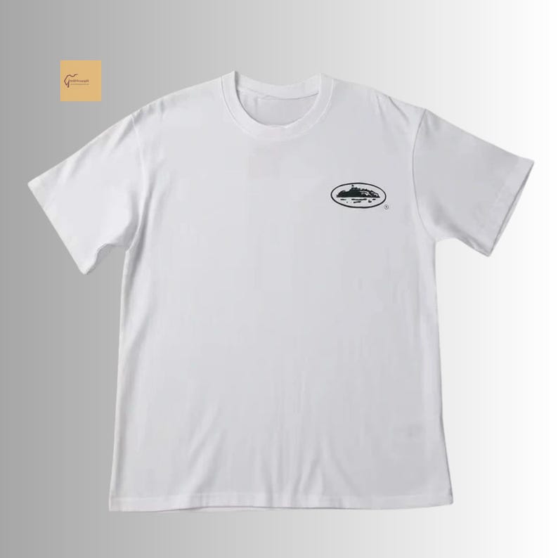 May include: A white short-sleeved t-shirt featuring a crew neckline and a small black oval graphic on the upper left chest. The graphic depicts a detailed landscape illustration inside the oval shape, positioned neatly against the plain white fabric.