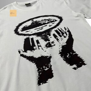 May include: A white short-sleeve t-shirt featuring a large black graphic print on the front. The design depicts two hands held upward, supporting a circular, textured shape that resembles a planet or a celestial body with a dark, grainy aesthetic.