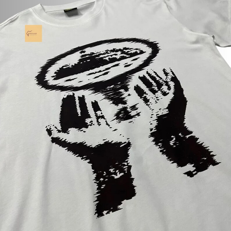 May include: A white short-sleeve t-shirt featuring a large black graphic print on the front. The design depicts two hands held upward, supporting a circular, textured shape that resembles a planet or a celestial body with a dark, grainy aesthetic.