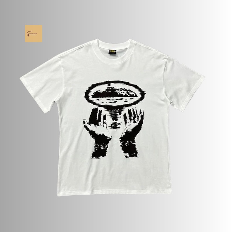 May include: A white short-sleeved t-shirt featuring a black graphic print on the chest. The design depicts two hands reaching upward toward an oval-shaped, textured island or landmass silhouette that appears to be floating above the palms.