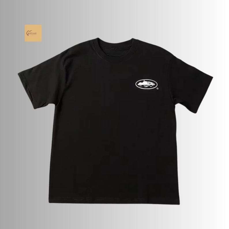 May include: A black short-sleeved t-shirt featuring a crew neckline and a small white oval graphic on the upper left chest. The graphic contains a white silhouette of a landscape or island shape inside the oval border, positioned on a plain background.