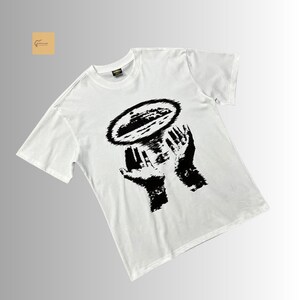 May include: A white short-sleeved t-shirt featuring a black graphic print on the chest. The design depicts two hands cupped upward, holding a circular, textured shape that resembles a landscape or island surrounded by a dark, radiating halo effect.