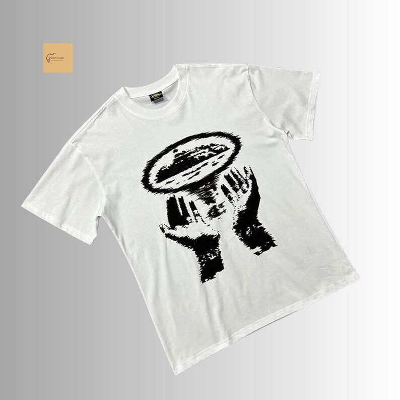 May include: A white short-sleeved t-shirt featuring a black graphic print on the chest. The design depicts two hands cupped upward, holding a circular, textured shape that resembles a landscape or island surrounded by a dark, radiating halo effect.