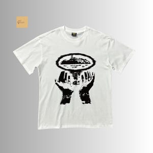 May include: A white short-sleeved t-shirt featuring a black graphic print on the chest. The design depicts two hands reaching upward toward an oval-shaped, textured island or landmass silhouette that appears to be floating above the palms.