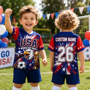 May include: A young child wearing a red, white, and blue USA soccer jersey and shorts. The jersey features an eagle, soccer ball, and the text "USA WORLD CUP 2026". The back of the jersey has the number 26 and the words "CUSTOM NAME".