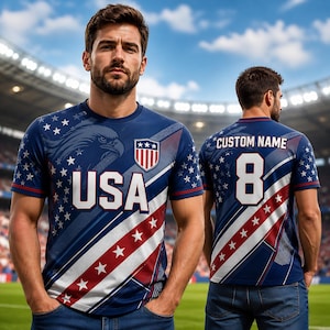 May include: A blue short-sleeved t-shirt featuring a graphic of a bald eagle, the text USA, and a shield emblem on the front. The back displays the text CUSTOM NAME and the number 8. The design includes white stars and diagonal red and white stripes.