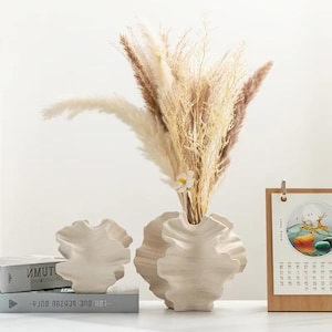 May include: A decorative arrangement featuring a cream-colored, ruffled vase holding dried pampas grass and a small white flower. A smaller matching vase and a calendar are also present. The arrangement is set on a white surface.