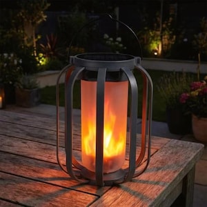 Puede incluir: Una linterna solar gris oscuro con asa, que presenta un efecto de llama realista. La linterna tiene un centro cilíndrico de vidrio esmerilado y está colocada sobre una mesa de madera. El efecto de llama emite una luz cálida naranja y amarilla.